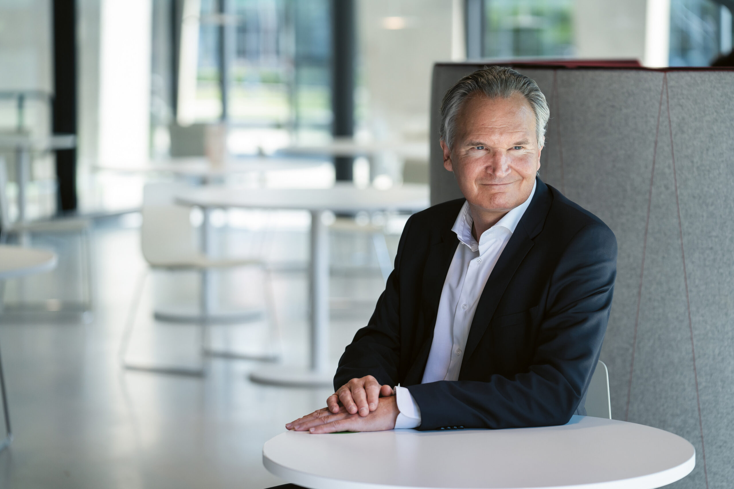 Ein Mann mittleren Alters in einem dunklen Anzug sitzt an einem runden weißen Tisch in einem modernen, hellen Büroraum, schaut zur Seite und lächelt leicht. Im Hintergrund sind leere weiße Stühle und Tische zu sehen.