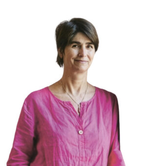 Frau mit kurzen braunen Haaren, die eine rosa Bluse und blaue Jeans trägt und lässig mit einer Hand in der Tasche vor einem weißen Hintergrund steht.