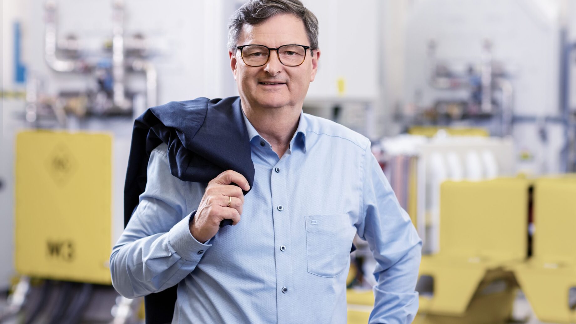 Ein Mann mittleren Alters mit Brille und grauen Haaren steht lächelnd in einem Haus, trägt ein hellblaues Button-up-Hemd und hält eine dunkle Jacke über die Schulter. Im unscharfen Hintergrund deuten Industrieanlagen auf Ortskernbelebung Steiermark hin.