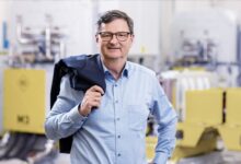 Ein Mann mittleren Alters mit Brille und grauen Haaren steht lächelnd in einem Haus, trägt ein hellblaues Button-up-Hemd und hält eine dunkle Jacke über die Schulter. Im unscharfen Hintergrund deuten Industrieanlagen auf Ortskernbelebung Steiermark hin.