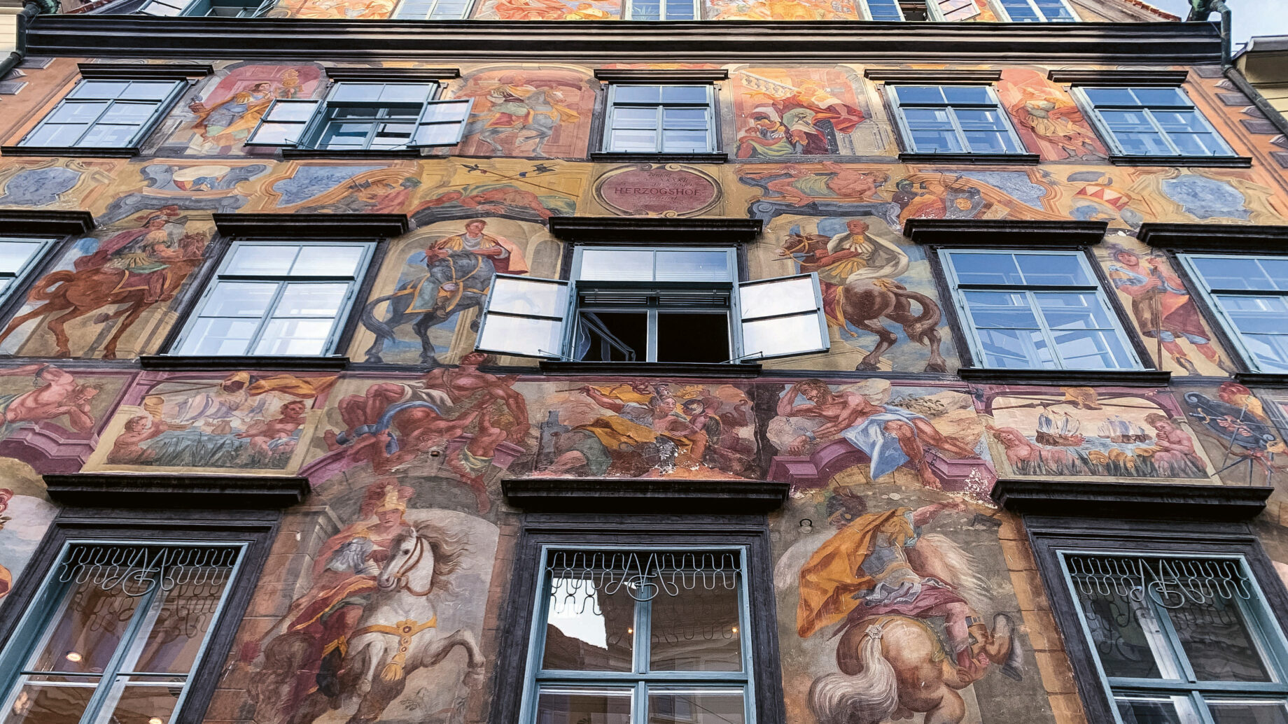 Eine Gebäudefassade mit bunten, detaillierten Fresken, die historische und mythische Szenen darstellen, mit mehreren Fenstern und einem offenen Fenster in der Mitte.