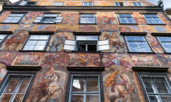 Eine Gebäudefassade mit bunten, detaillierten Fresken, die historische und mythische Szenen darstellen, mit mehreren Fenstern und einem offenen Fenster in der Mitte.
