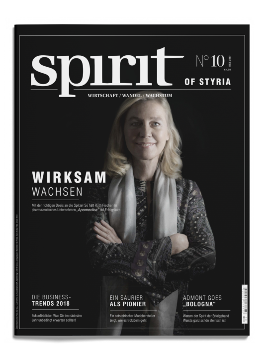 Titelbild der Zeitschrift "spirit of styria" mit einer lächelnden Frau mit blondem Haar und verschränkten Armen, die einen gemusterten Schal und ein dunkles Oberteil trägt.