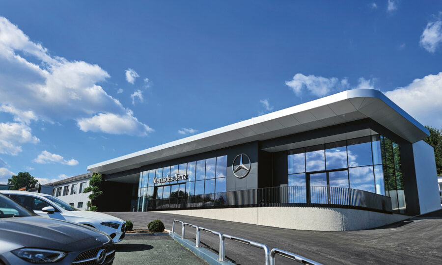 Modernes Gebäude eines Mercedes-Benz-Händlers mit großen Fenstern, einem auffälligen Logo und geparkten Autos davor unter einem strahlend blauen Himmel mit vereinzelten Wolken.