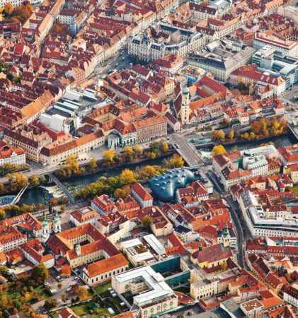 Luftaufnahme einer Stadt mit roten Dächern, einem Fluss, Brücken und herbstlichen Bäumen, die über die Stadtlandschaft verstreut sind.