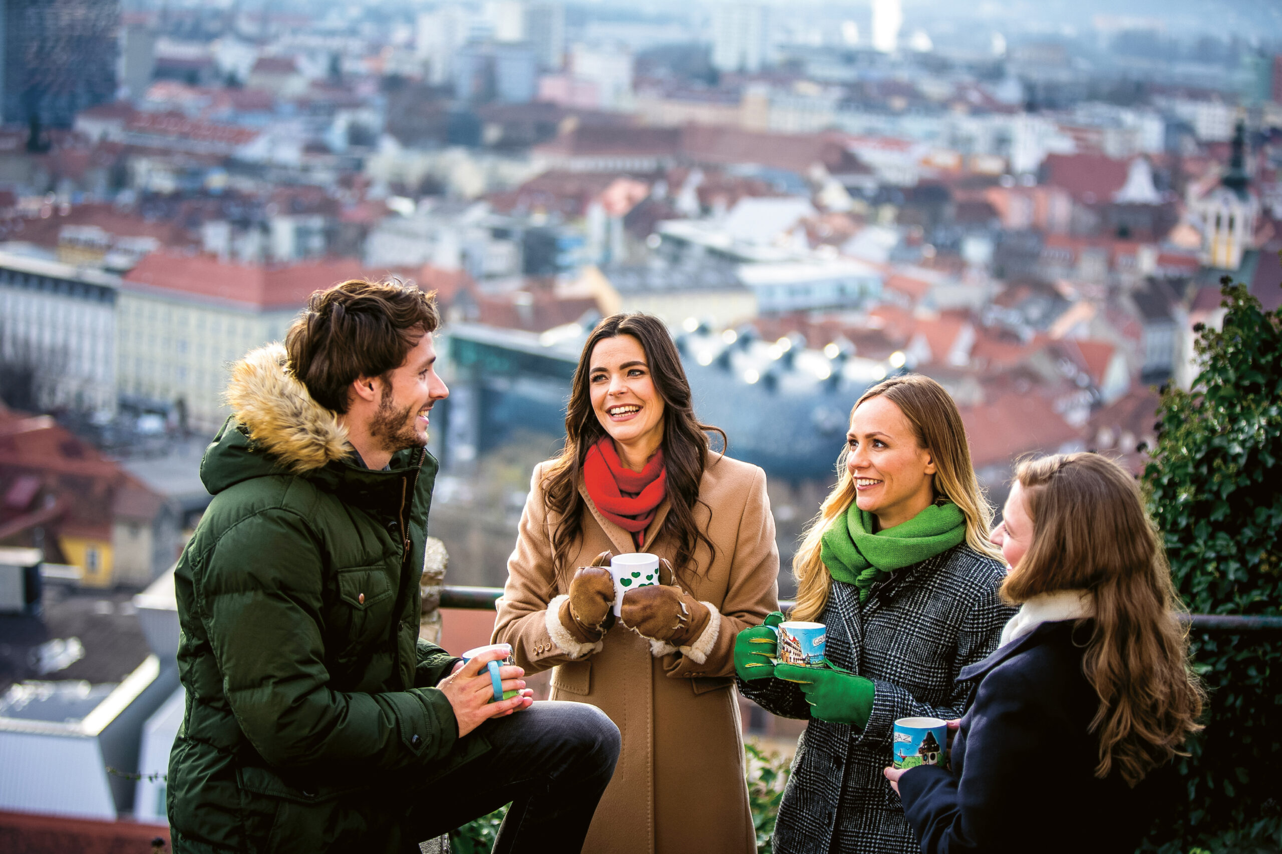 Vier lächelnde Menschen in Winterkleidung stehen im Freien auf einem Hügel, halten Tassen in der Hand und haben eine Stadtlandschaft im Hintergrund.