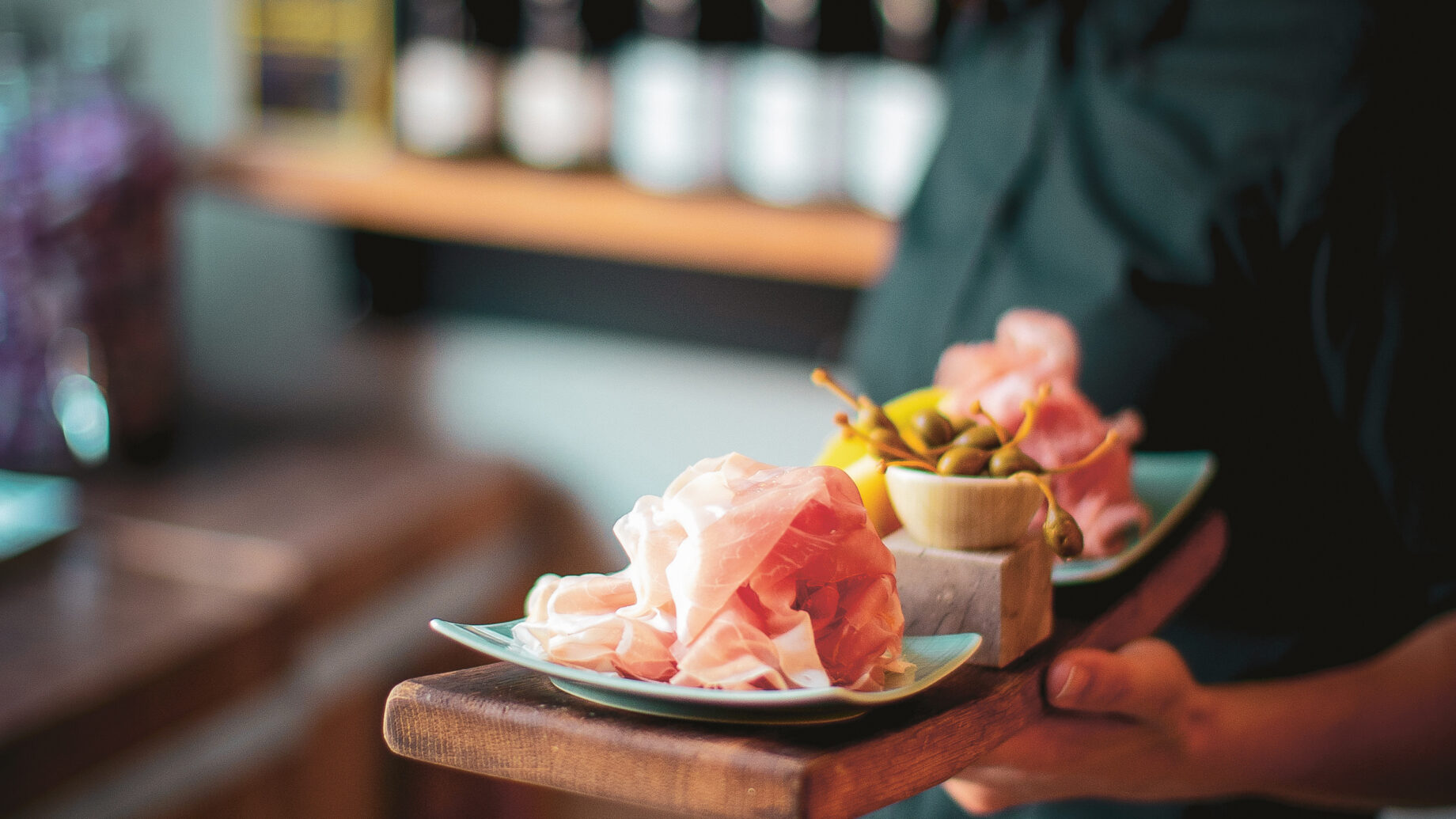 Ein schwarz gekleideter Kellner hält in einem Restaurant ein Holztablett mit Wurstwaren und Oliven, im Hintergrund sind unscharfe Flaschen zu sehen.