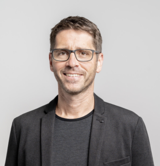 Mann mit kurzen braunen Haaren und Brille, der einen schwarzen Blazer und ein dunkles Hemd trägt, lächelt vor einem schlichten hellgrauen Hintergrund.