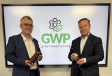 Zwei Männer in Anzügen stehen vor einem Bildschirm mit dem Logo von Greenwood Power und halten jeweils ein Elektrogerät der Herbert Temmel GmbH Steiermark in der Hand.