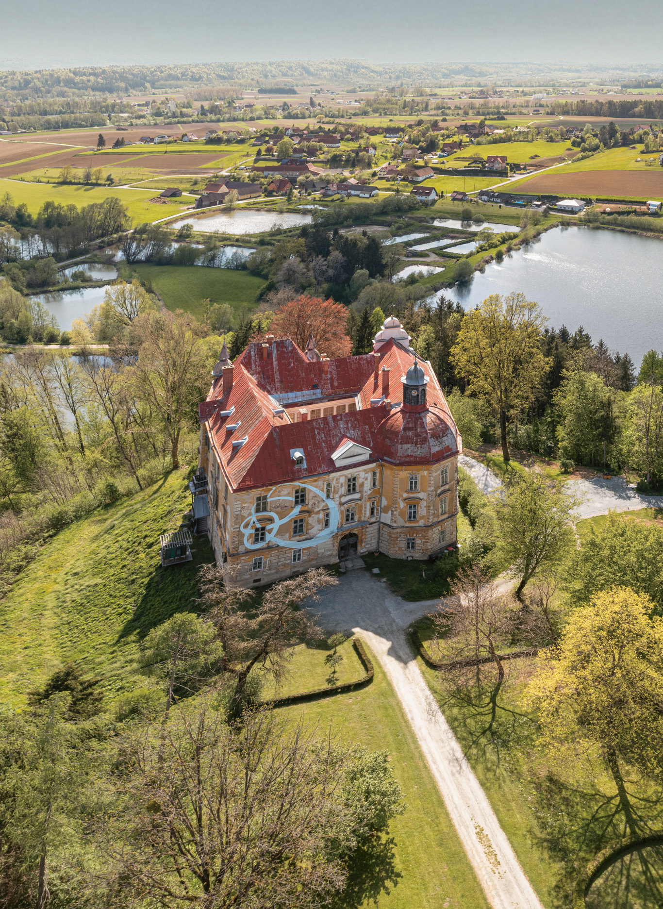 Luftaufnahme eines historischen Backsteinhauses mit rotem Dach, umgeben von Bäumen, Teichen und grünen Feldern unter einem klaren Himmel.