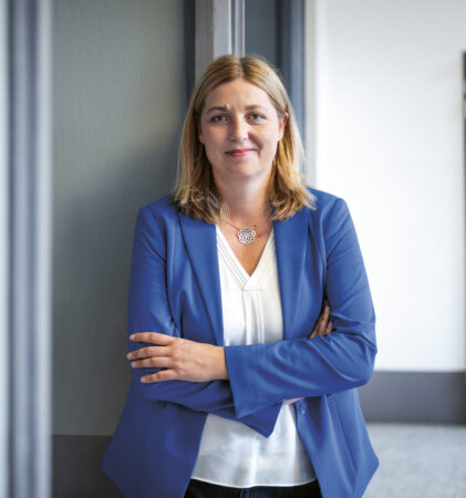 Eine Frau mit blondem Haar, blauem Blazer und weißer Bluse steht in einem Innenraum und lächelt mit verschränkten Armen gegen eine graue Wand.