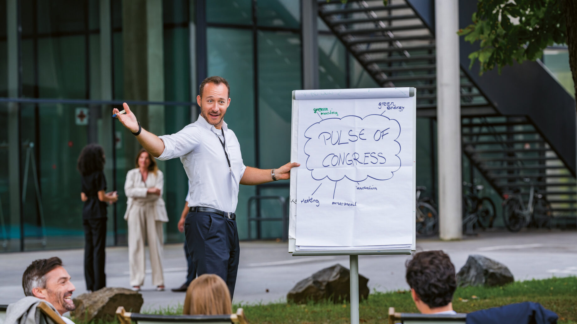 Ein Mann hält einen Vortrag im Freien vor einer sitzenden Gruppe, wobei er auf ein Flipchart mit der Aufschrift "Pulse of Congress" zeigt, mit einem modernen Gebäude im Hintergrund.