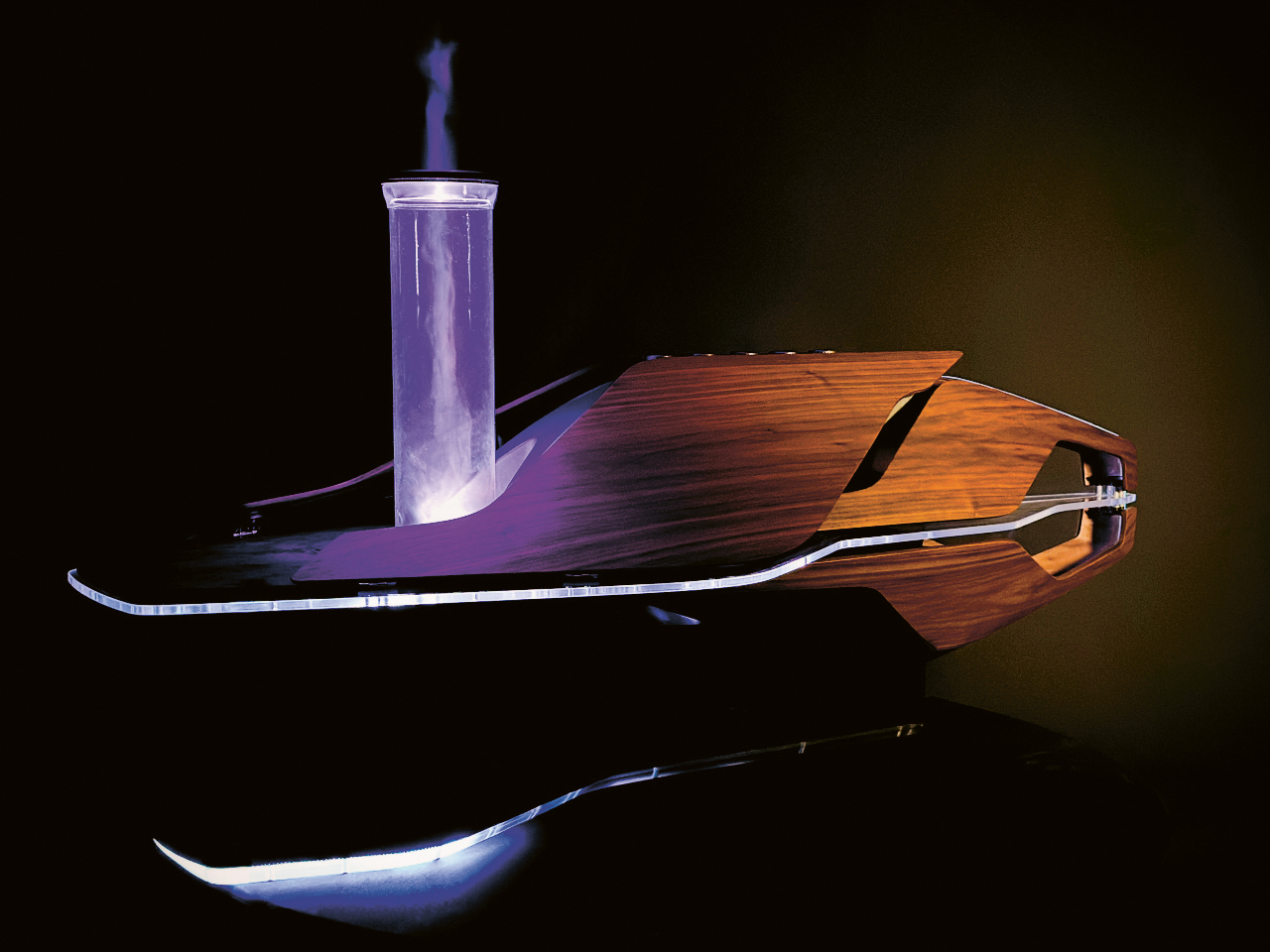 Ein schlankes, modernes Holzgerät stößt vor einem dunklen Hintergrund eine violett leuchtende Rauchsäule aus, deren Spiegelung darunter sichtbar ist.