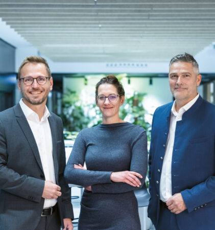 Drei professionell gekleidete Personen stehen in einem modernen Büro, lächeln und schauen in die Kamera, während im Hintergrund Grünflächen zu sehen sind.