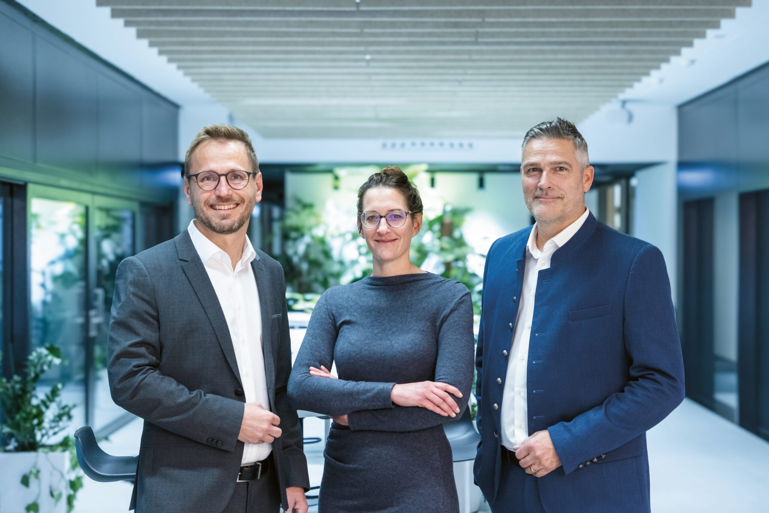 Drei professionell gekleidete Personen stehen in einem modernen Büro, lächeln und schauen in die Kamera, während im Hintergrund Grünflächen zu sehen sind.