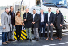 Eine Gruppe von neun Personen in Geschäftskleidung steht lächelnd vor einem Lastwagen und einer Tankstelle, als Emerald Horizon Robert Holzmann zum Vizepräsidenten ernennt.