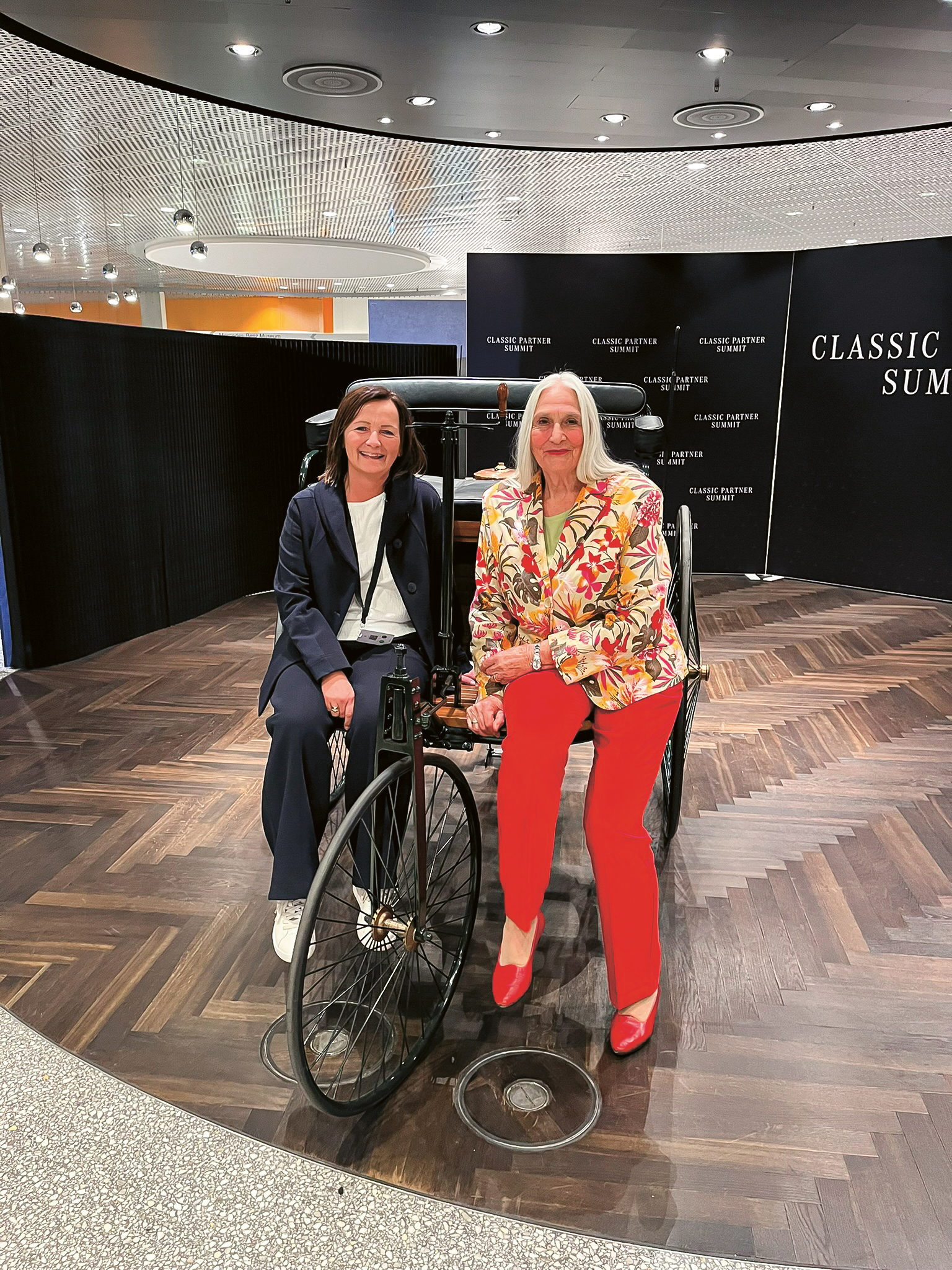 Zwei Frauen posieren und lächeln zusammen in einem Innenraum, auf einem altmodischen Dreirad sitzend, vor einem Display mit der Aufschrift CLASSIC FUTURE SUMMIT".