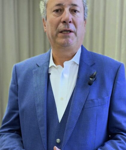 Ein Mann im blauen Anzug wird in einem Innenraum interviewt, die Hände gefaltet, mit einem Mikrofon.