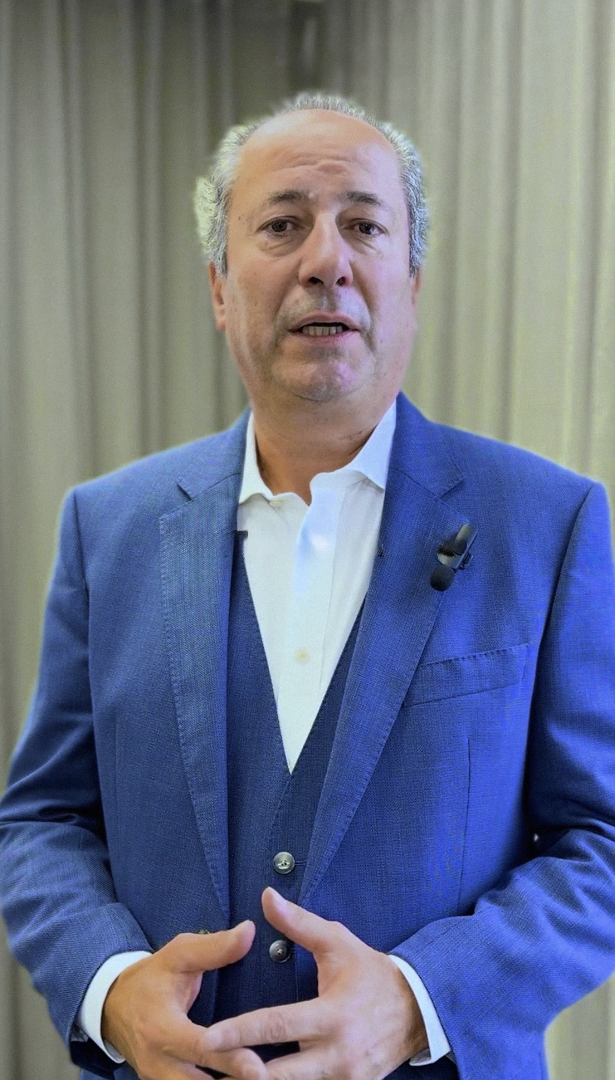 Ein Mann im blauen Anzug wird in einem Innenraum interviewt, die Hände gefaltet, mit einem Mikrofon.