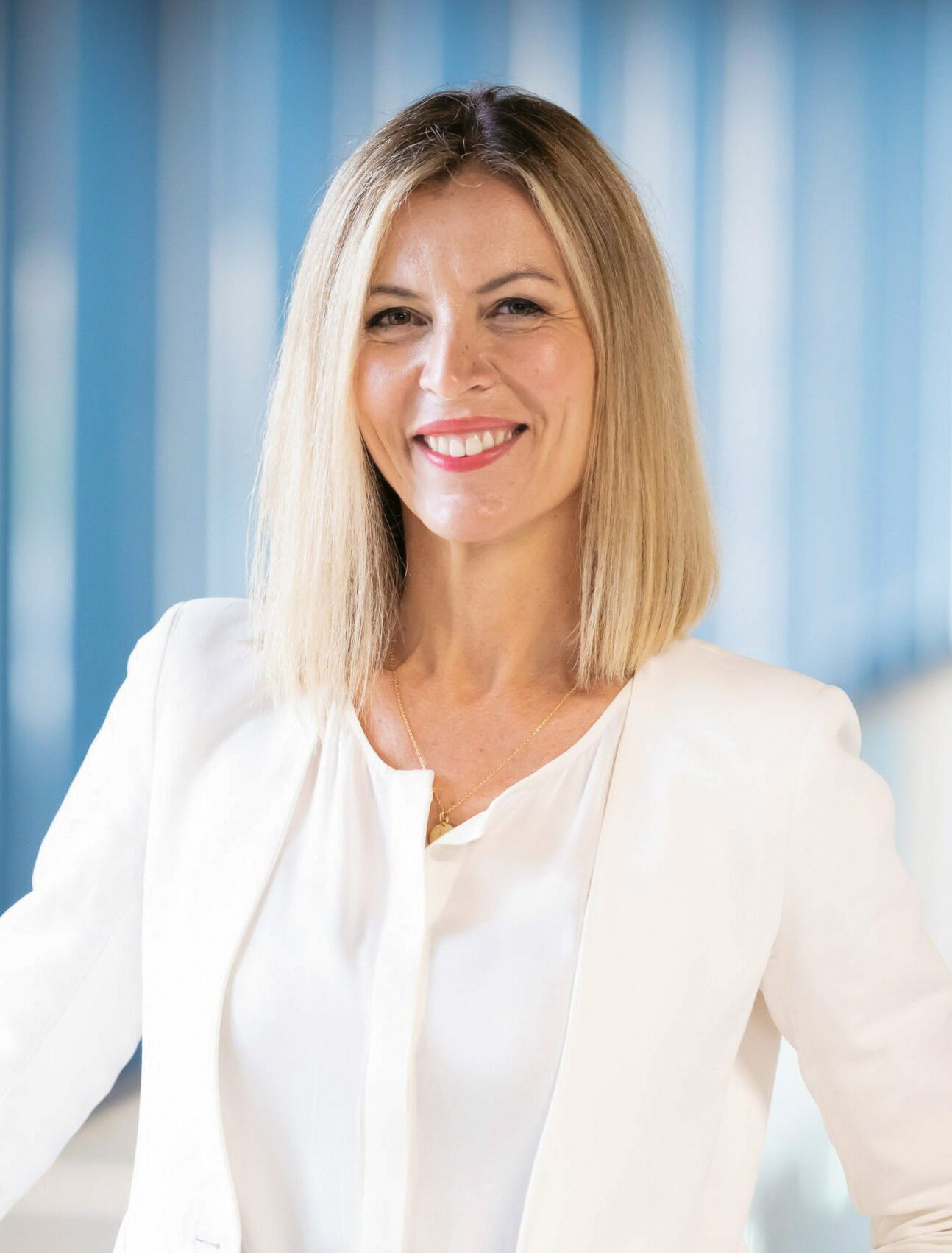 Eine lächelnde Frau mit glattem blondem Haar, die einen weißen Blazer und eine weiße Bluse trägt, steht in einem hell erleuchteten modernen Flur.