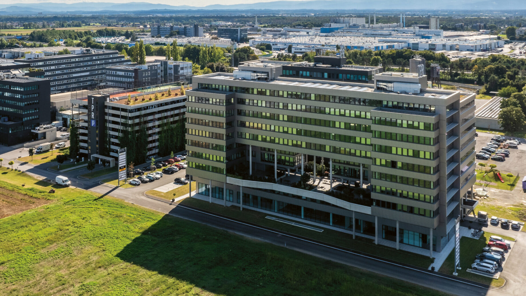 Luftaufnahme von modernen Bürogebäuden und Parkplätzen neben grasbewachsenen Feldern unter einem teilweise bewölkten Himmel.