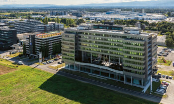 Luftaufnahme von modernen Bürogebäuden und Parkplätzen neben grasbewachsenen Feldern unter einem teilweise bewölkten Himmel.