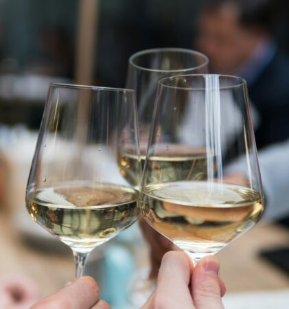 Nahaufnahme von drei Personen, die gemeinsam auf ein Glas Weißwein anstoßen, mit einem unscharfen Hintergrund von Menschen, die an einem Tisch sitzen.