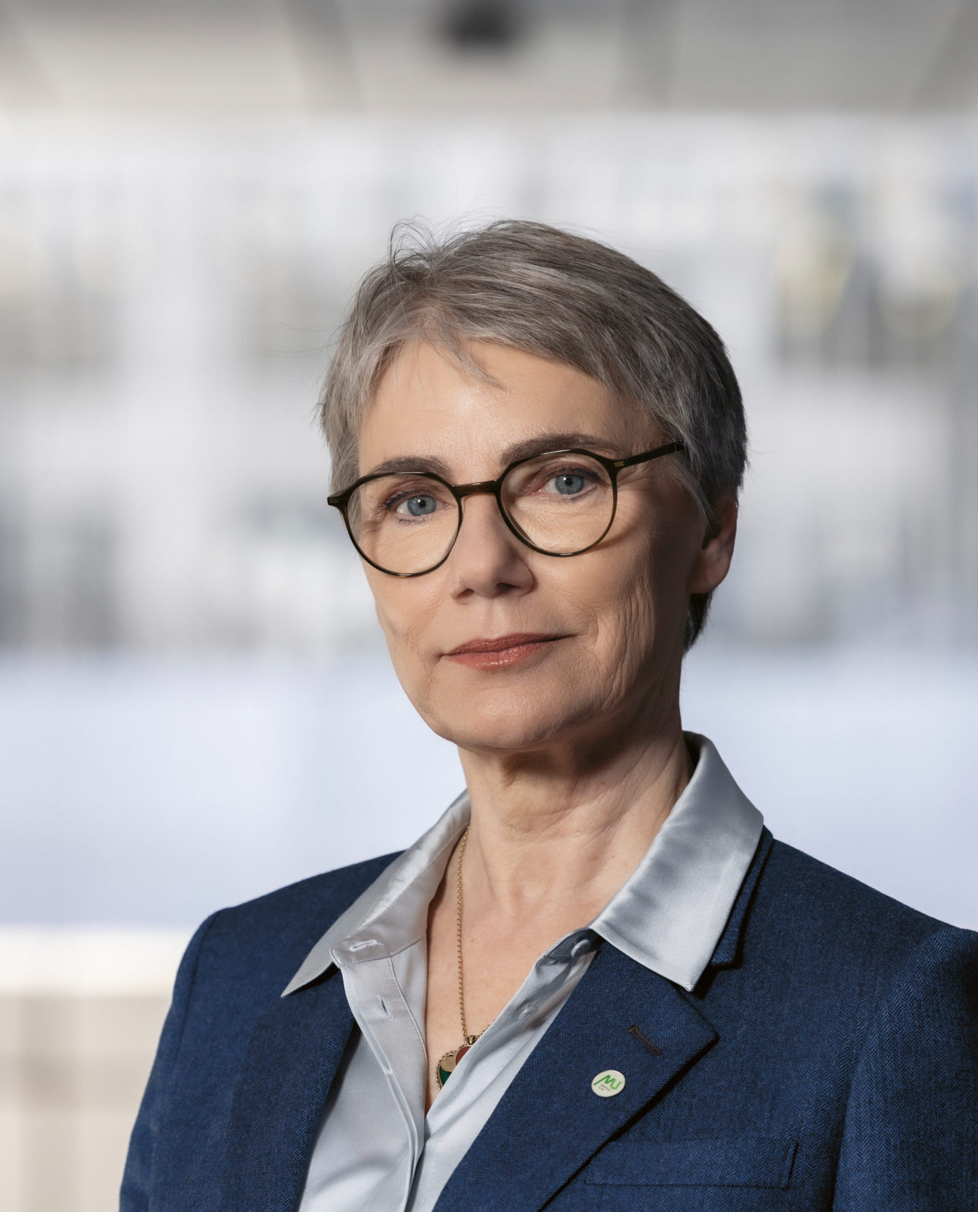 Eine ältere Frau mit kurzen grauen Haaren und Brille trägt einen blauen Blazer und eine silberne Bluse und steht in einer hellen, modernen Büroumgebung.