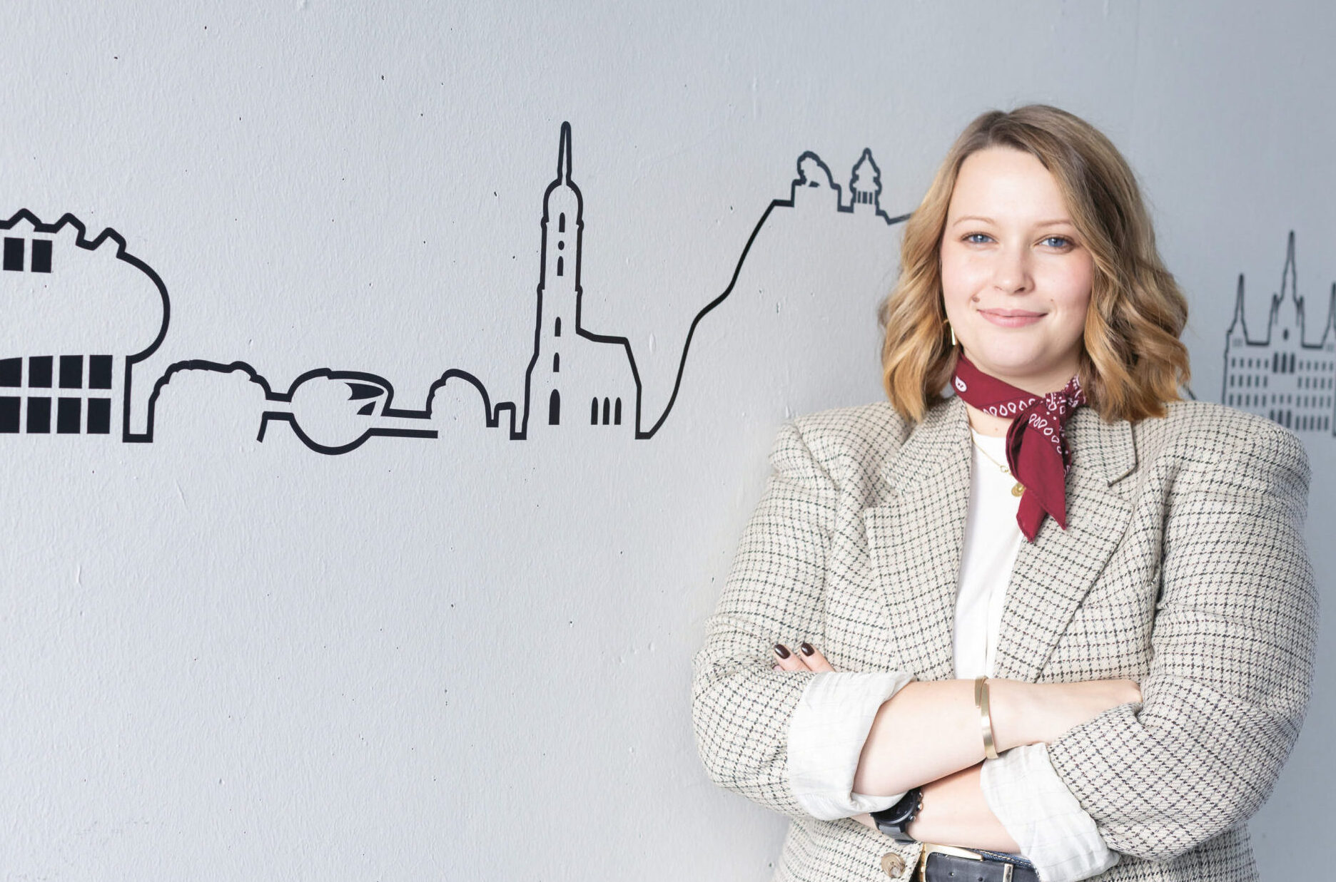 Eine Frau mit blondem Haar, die einen karierten Blazer und einen roten Schal trägt, steht lächelnd mit verschränkten Armen vor einer Wand mit einer Abbildung der Skyline einer Stadt.