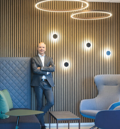 Ein Mann im Anzug steht lächelnd mit verschränkten Armen in einer modernen Lounge mit holzgetäfelten Wänden, kreisrunden Leuchten und modernen Möbeln.