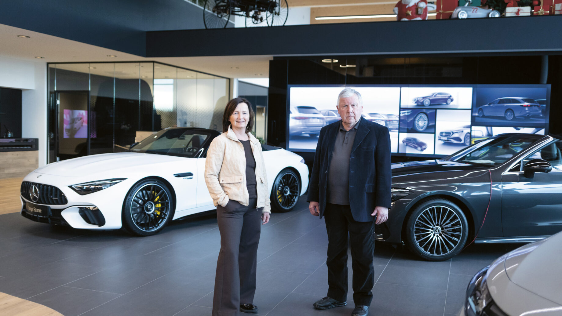 Zwei Personen stehen in einem modernen Autohaus mit Luxusfahrzeugen, darunter ein weißes Cabrio und eine graue Limousine, die im Hintergrund ausgestellt sind.