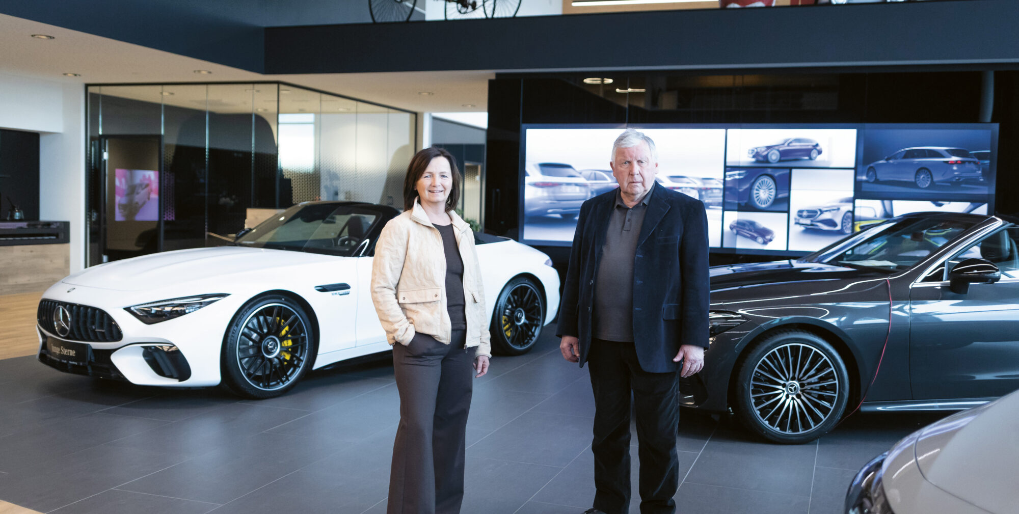 Zwei Personen stehen in einem modernen Autohaus mit Luxusfahrzeugen, darunter ein weißes Cabrio und eine graue Limousine, die im Hintergrund ausgestellt sind.