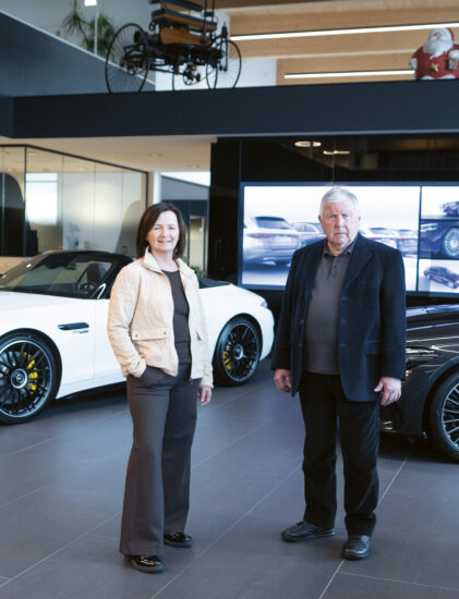 Zwei Personen stehen in einem modernen Autohaus mit Luxusfahrzeugen, darunter ein weißes Cabrio und eine graue Limousine, die im Hintergrund ausgestellt sind.