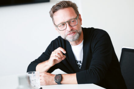 Ein Mann mit Brille und Bart sitzt an einem Tisch, gestikuliert mit einer Hand, lächelt und hat ein Glas Wasser vor sich stehen.