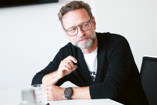Ein Mann mit Brille und Bart sitzt an einem Tisch, gestikuliert mit einer Hand, lächelt und hat ein Glas Wasser vor sich stehen.