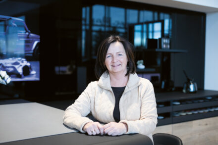 Eine Frau mit dunklem Haar und einer hellen Jacke steht lächelnd in einer modernen Küche mit dunklen Schränken und einer reflektierenden Aufkantung.