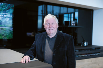 Ein älterer Mann mit weißem Haar, der einen dunklen Blazer und ein Polohemd trägt, steht lächelnd in einer modernen Kücheneinrichtung.