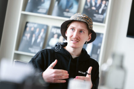 Eine Person mit einem Eimerhut mit Leopardenmuster und einem schwarzen Kapuzenpulli gestikuliert, während sie spricht, während im Hintergrund verschwommene Zeitschriften in Regalen ausliegen.