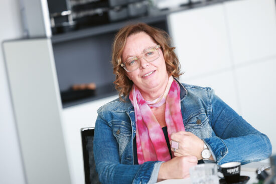 Eine Frau mit Brille und rosa Schal lächelt, während sie an einem Tisch sitzt und eine Jeansjacke in einem modernen Innenraum trägt.