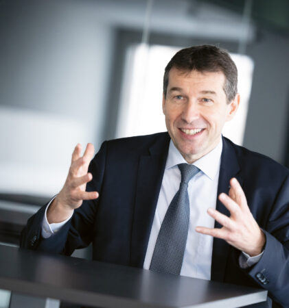 Ein Mann im Anzug gestikuliert mit seinen Händen und lächelt, während er an einem Tisch in einem modernen Büro sitzt.