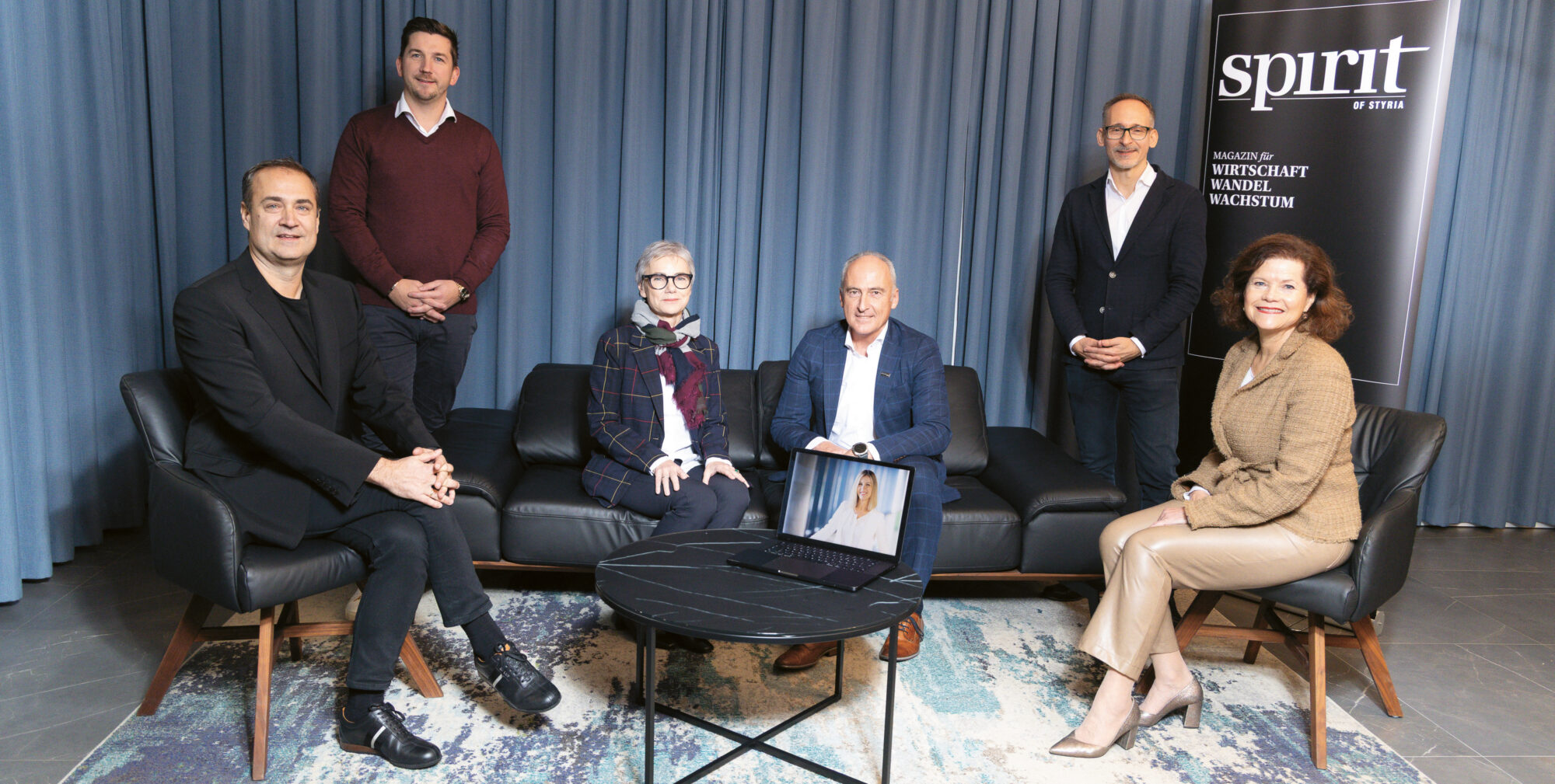 Sechs Personen, vier sitzend und zwei stehend, posieren in einem modernen Raum mit blauen Vorhängen, einem Teppich und einem "Spirit of Styria"-Banner; auf dem Tisch steht ein Laptop.