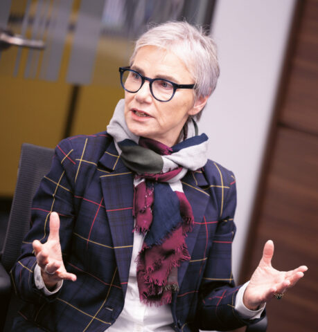 Ältere Frau mit kurzen grauen Haaren, Brille und einem karierten Blazer spricht angeregt und gestikuliert mit beiden Händen, während sie in einem Haus sitzt.