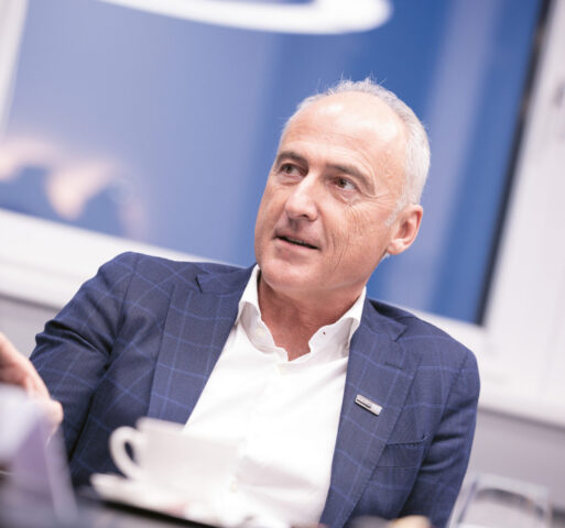 Ein Mann in einem blaukarierten Anzug sitzt an einem Tisch mit einer Kaffeetasse, spricht und gestikuliert mit der Hand in einer hellen Büroumgebung.