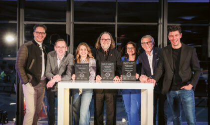 Sieben Personen stehen lächelnd hinter einem Tisch mit Urkunden und posieren gemeinsam in einem gut beleuchteten Raum mit großen Fenstern im Hintergrund.