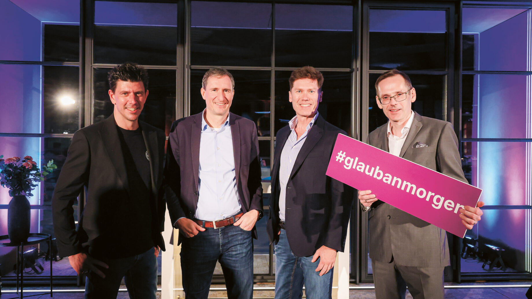 Vier Männer in legerer Business-Kleidung stehen in einem Innenraum zusammen; einer hält ein Schild mit der Aufschrift "#glaubanmorgen", das lila beleuchtet ist.