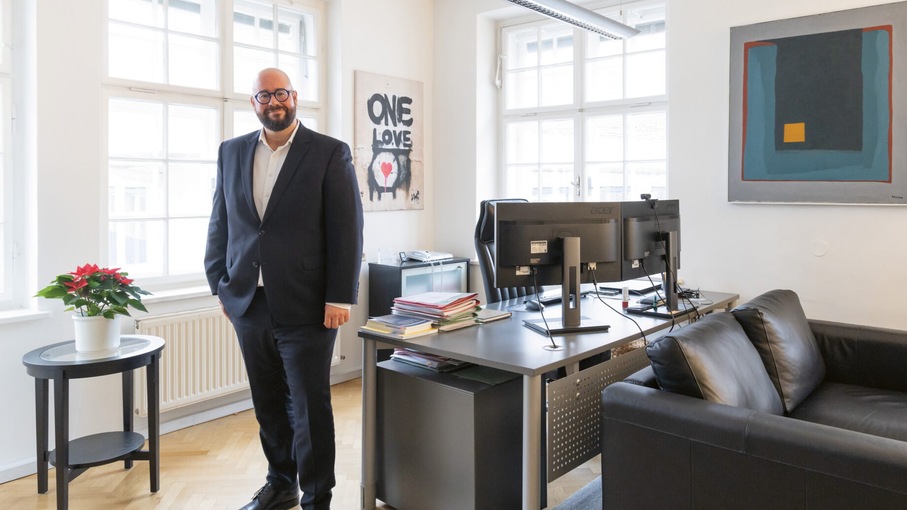 Ein Mann im Anzug steht lächelnd in einem modernen Büro mit großen Fenstern, einem Schreibtisch, einer schwarzen Couch und Kunstwerken an den Wänden.