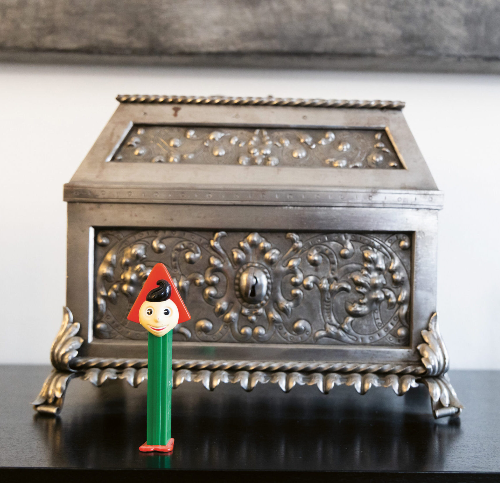 Ein bunter Pinocchio-PEZ-Spender steht vor einer verzierten silbernen Schachtel und einem beigen Gösser-Becher auf einer schwarzen Fläche.