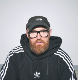 Ein bärtiger Mann mit Brille und Adidas-Trainingsanzug sitzt im Schneidersitz auf dem Boden in einem leeren, weißwandigen Raum, der von oben beleuchtet wird.