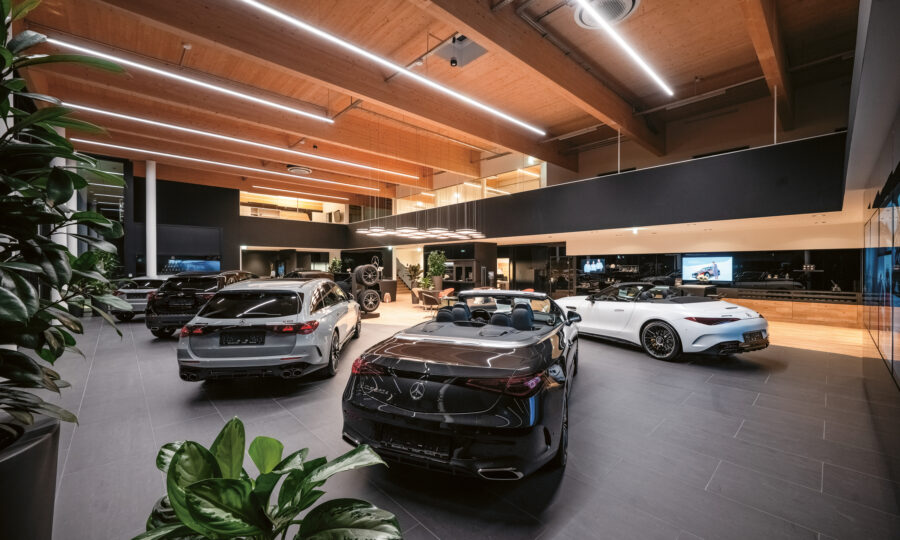 Modernes Autohaus mit ausgestellten Luxusfahrzeugen, mit eleganter Beleuchtung, hohen Decken, großen Fenstern und Grünpflanzen im Vordergrund.