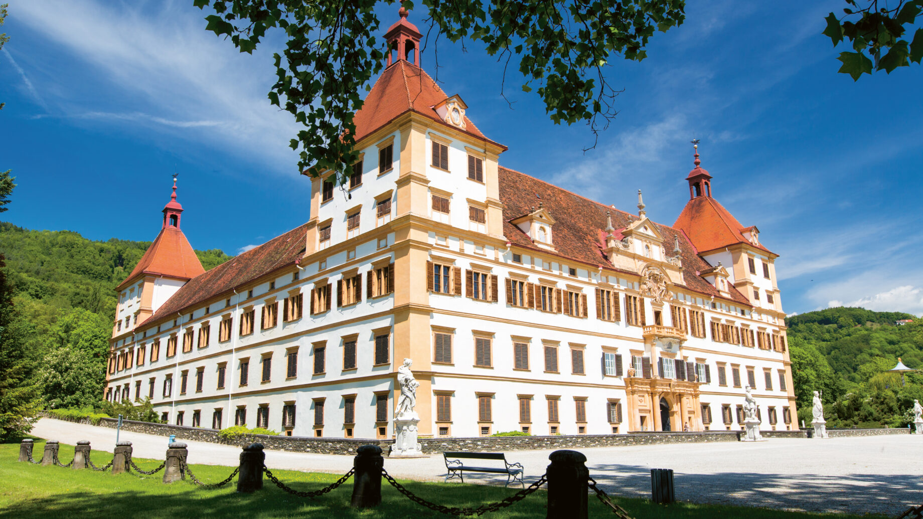 Ein großer, reich verzierter Barockpalast mit roten Dächern und weißen Wänden steht unter blauem Himmel, umgeben von Grün und Statuen.
